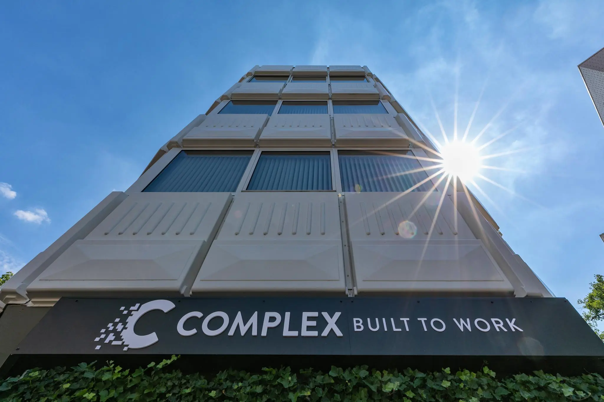 Vooraanzicht van het moderne kantoorgebouw COMPLEX aan de 1e Hogeweg, met de zon die schijnt aan een heldere blauwe lucht.